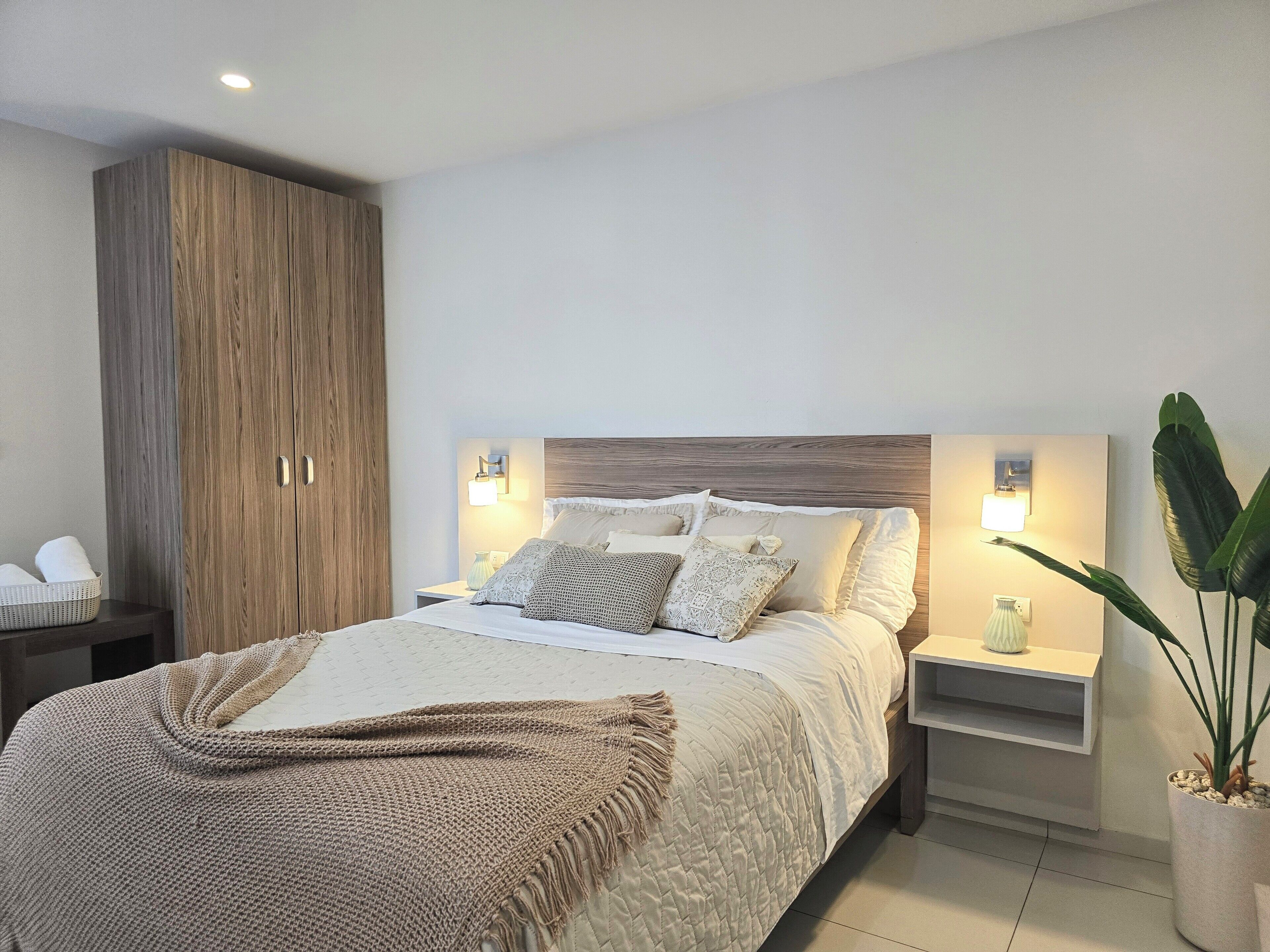 Quarto moderno com cama grande, guarda-roupa de madeira, abajures e um vaso de planta.
