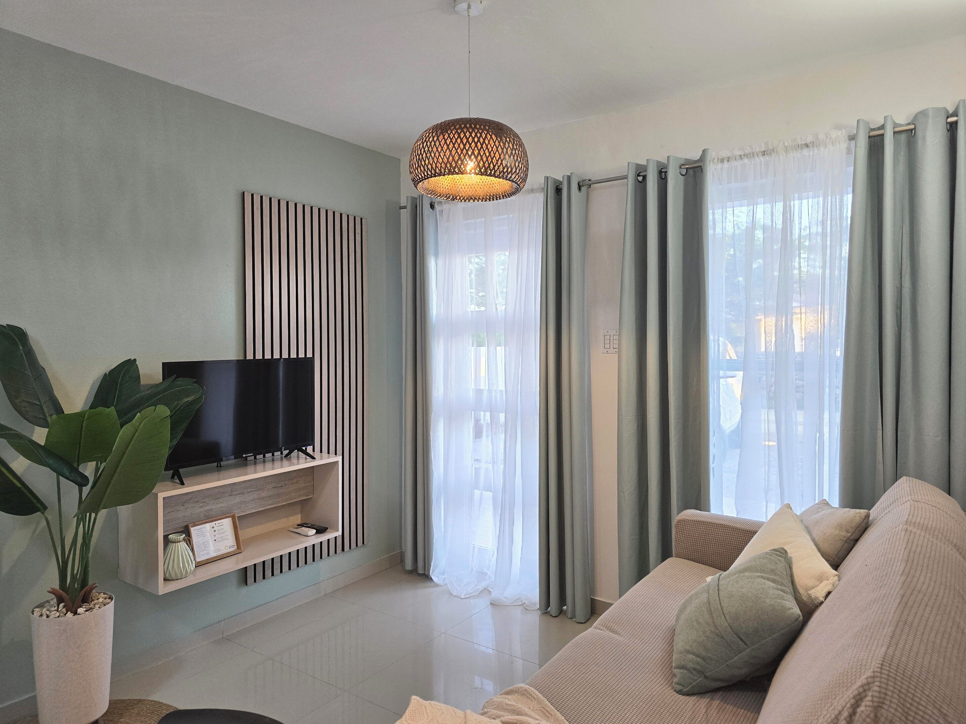Een moderne woonkamer met een flatscreen televisie op een houten standaard, een grote potplant, een beige bank met kussens en een hanglamp.
