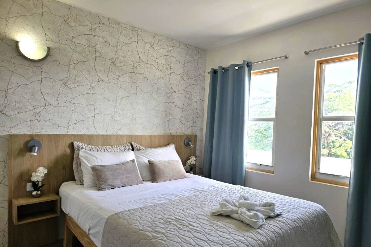 Un dormitorio con cabecera de madera, colcha blanca acolchada, dos mesitas de noche con lámparas y vistas a la vegetación a través de dos ventanas.
