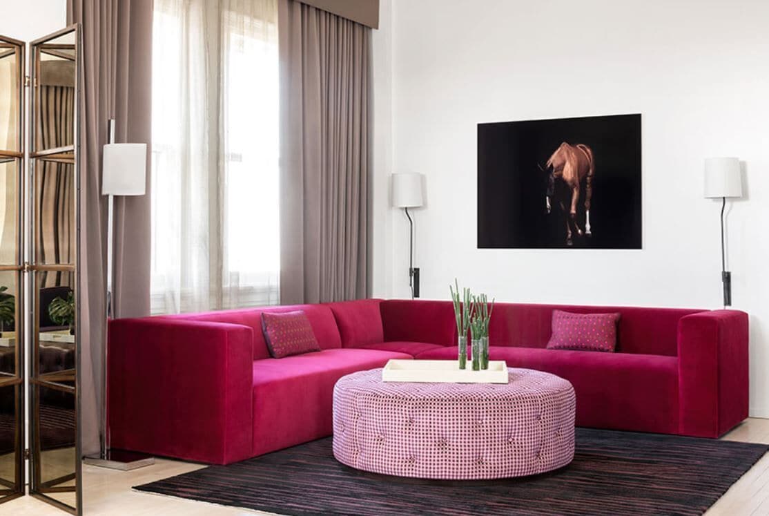 Una sala de estar moderna con un sofá seccional rojo intenso, un otoman redondo con funda a cuadros y un cuadro de un caballo en blanco y negro en la pared.
