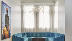 Een moderne woonkamer met een blauwe hoekbank, een ronde salontafel en een groot raam met transparante gordijnen en een decoratieve kroonluchter.