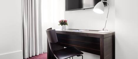 A modern hotel room with a dark wood desk, a black chair, a white desk lamp, a flat-screen TV, and a vase with flowers.