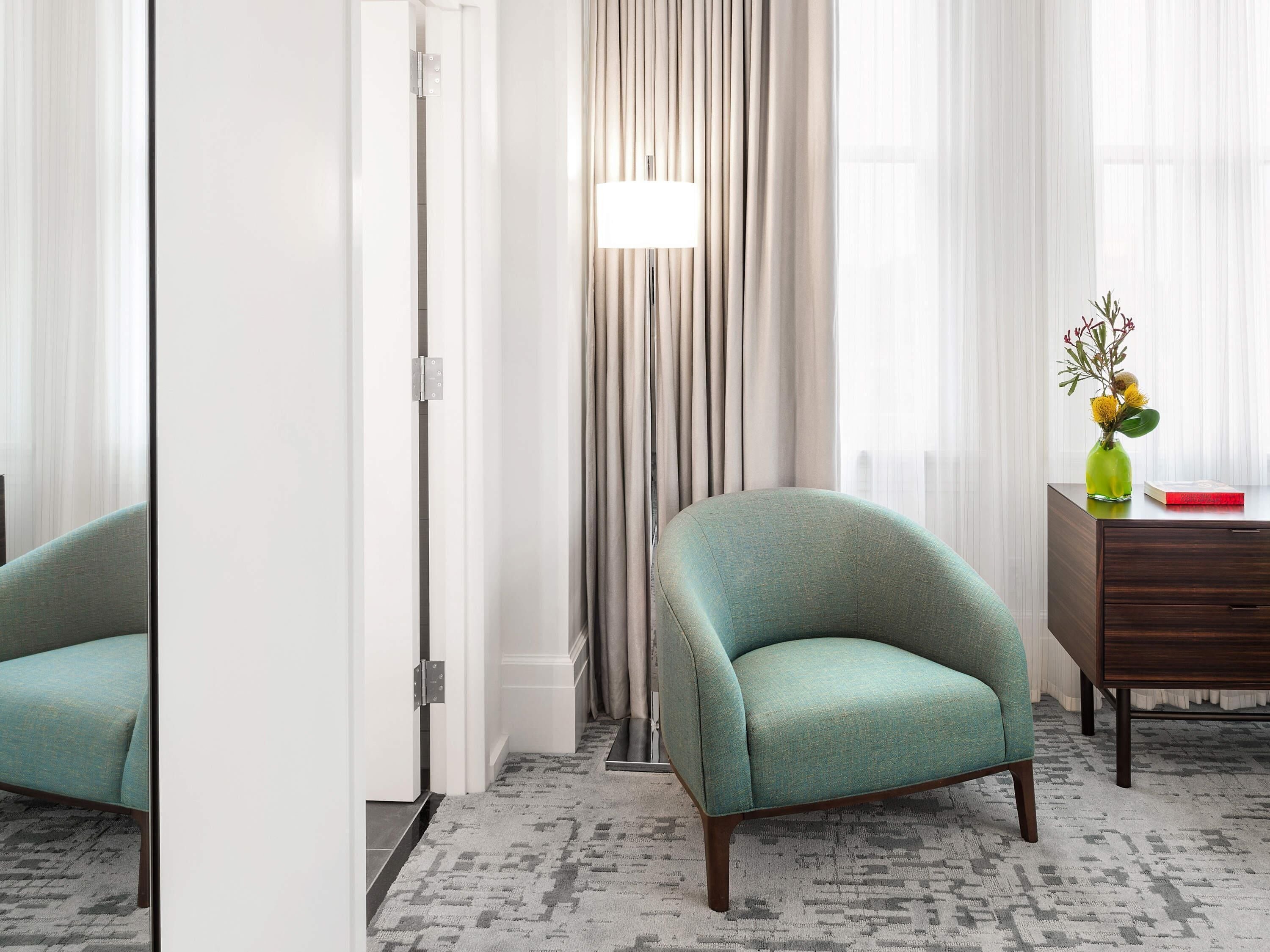 A room with a teal armchair, a wooden dresser, a floor lamp, and a vase with flowers.