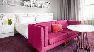 A modern hotel room with a white bed, a pink sofa, a round white table, and a wall-mounted artwork of horses.
