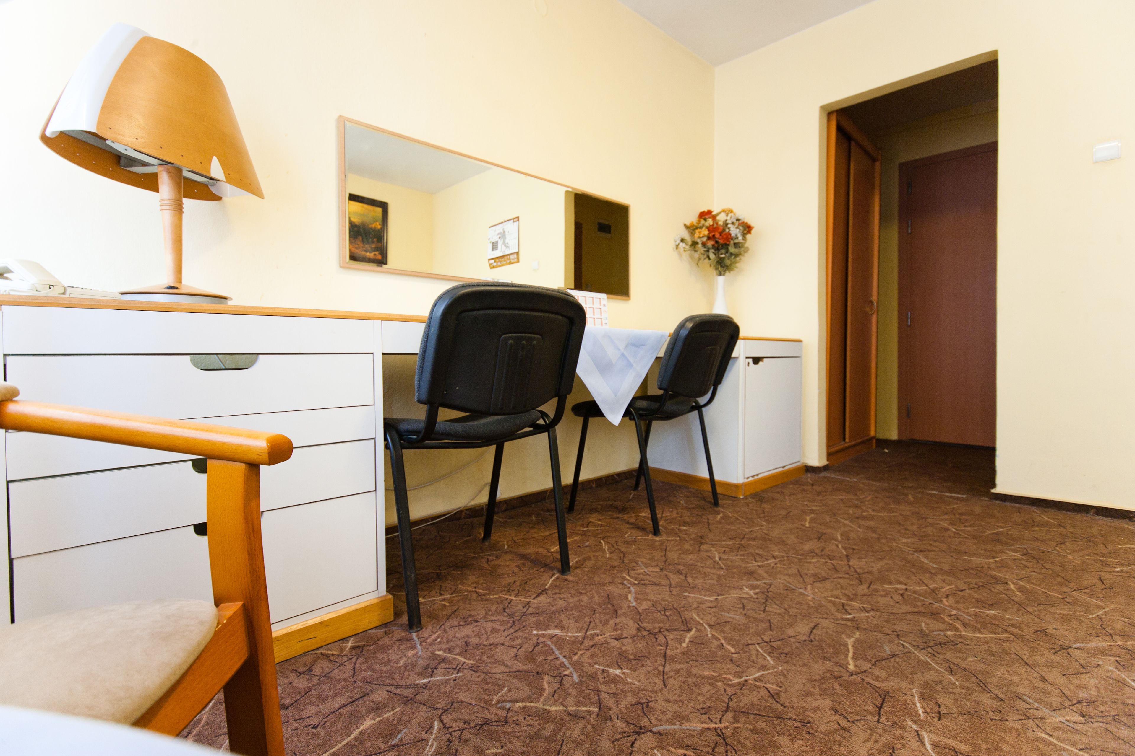 A hotel room with a desk, two chairs, a lamp, a dresser, and a door.