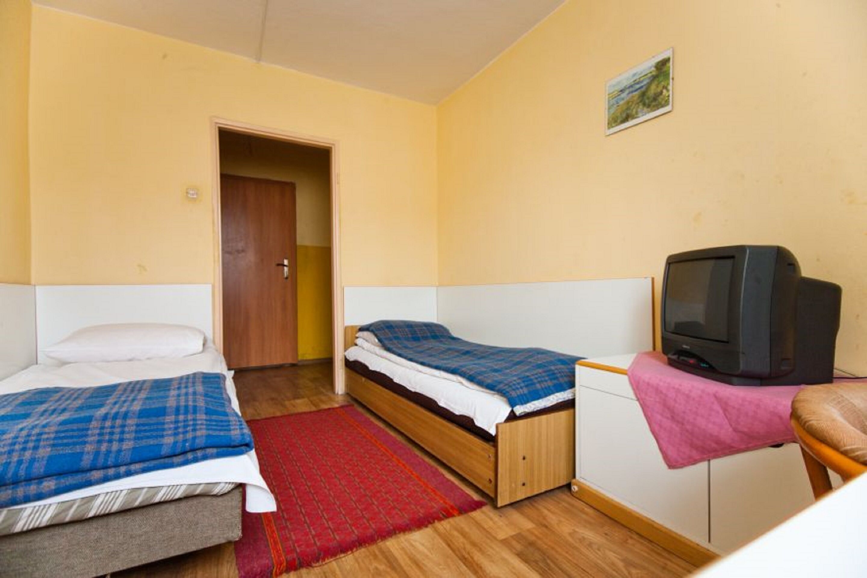 Una habitación pequeña con dos camas individuales, un televisor sobre un soporte y una puerta que da a otra habitación.