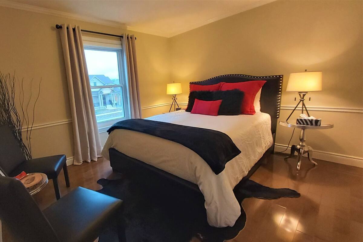Habitación de hotel con cama, dos lámparas de noche, una silla, una mesita y vistas al exterior a través de una ventana con cortinas.