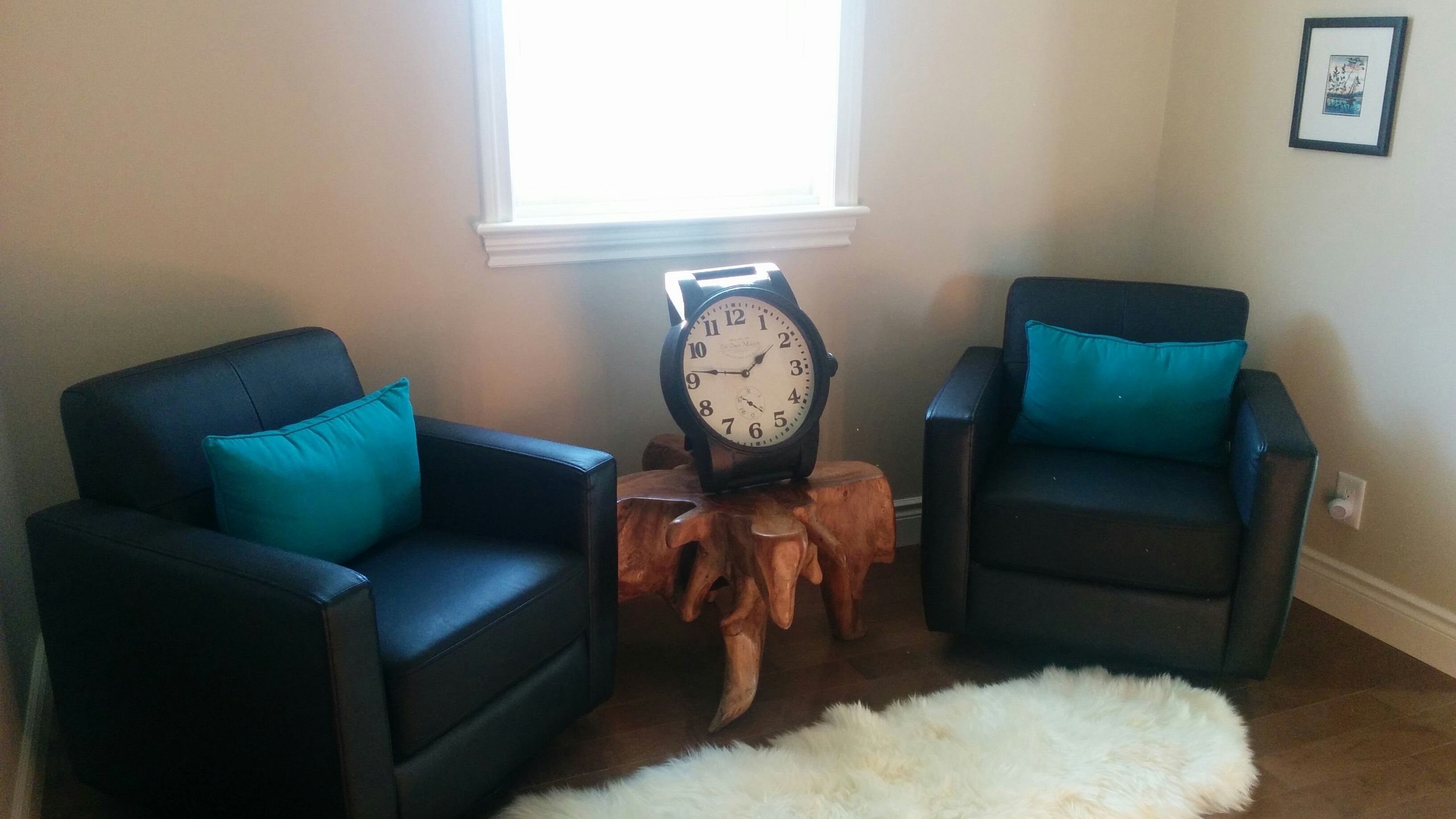 Sala de estar con dos sillones negros con cojines color turquesa, una mesa de centro de madera y una alfombra mullida blanca. Se ve un reloj de pared.