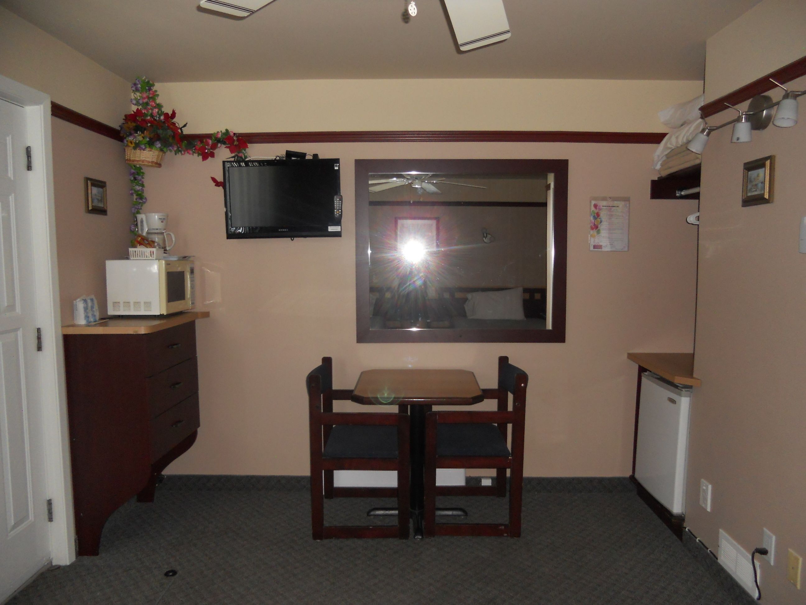 Una kitchenette pequeña con microondas, cafetera y lavamanos. Hay una mesa de madera y dos sillas. Un televisor está montado en la pared sobre la mesa.