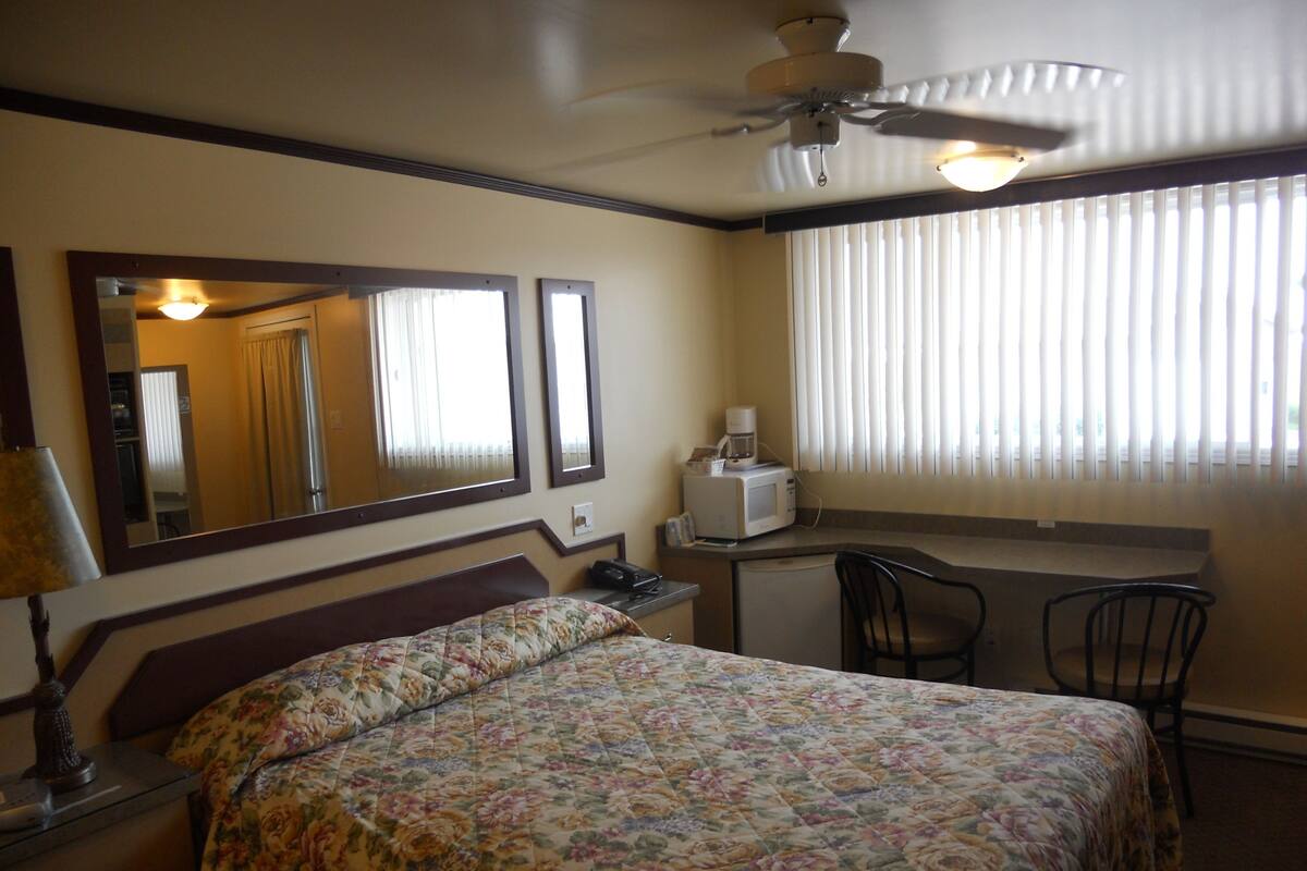Habitación de hotel con cama, escritorio, silla, ventilador de techo y ventana con persianas.