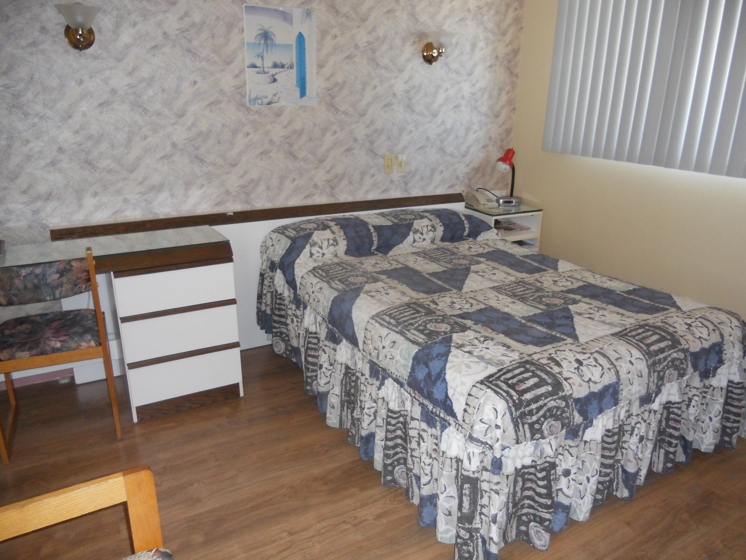 Un dormitorio con una cama de madera, un escritorio con silla, una mesita de noche y una lámpara de pared.