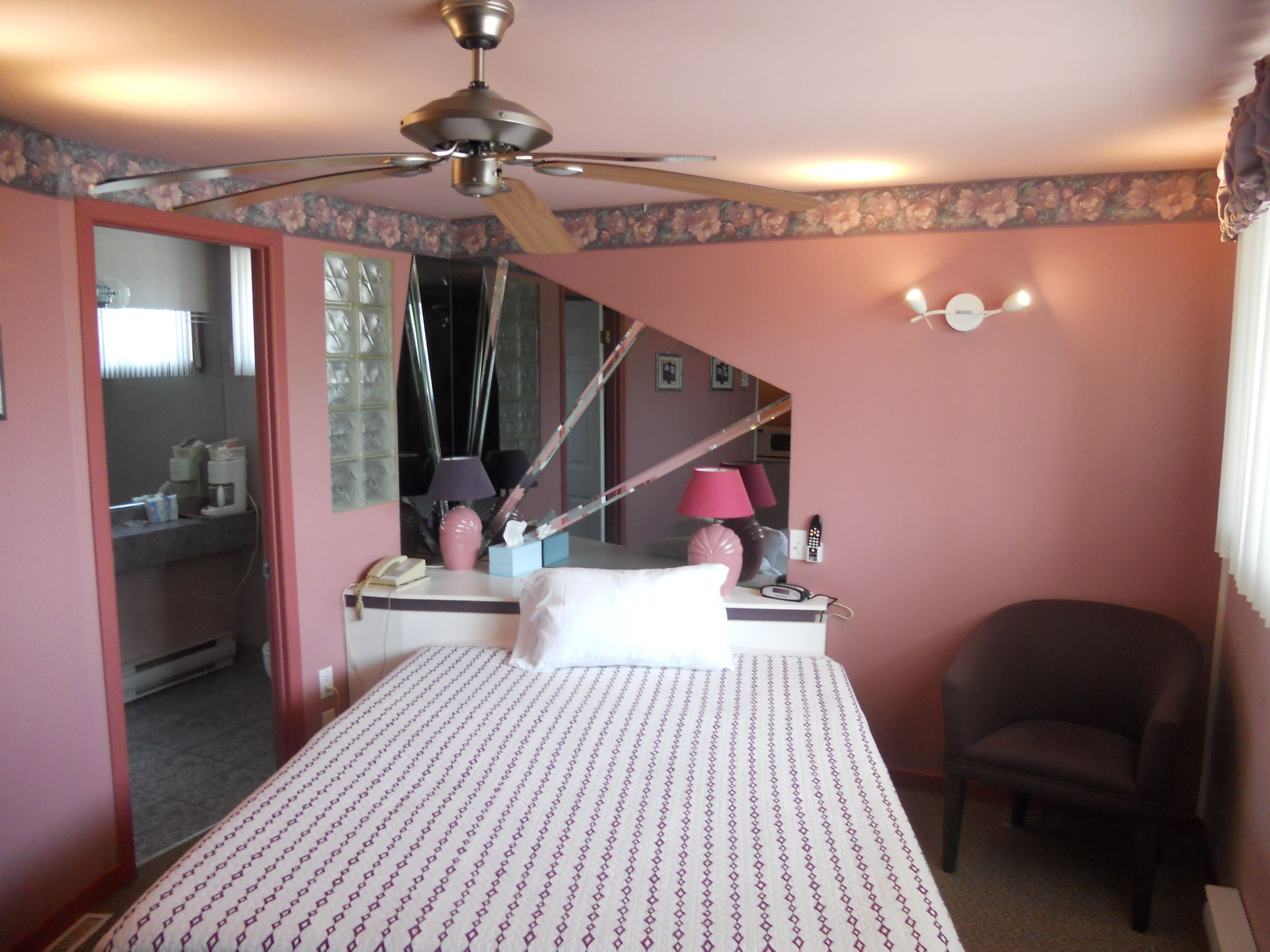 A bedroom with a bed, a chair, a desk with a lamp, a telephone, and a ceiling fan.