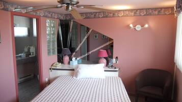A bedroom with a bed, a chair, a desk with a lamp, a telephone, and a ceiling fan.