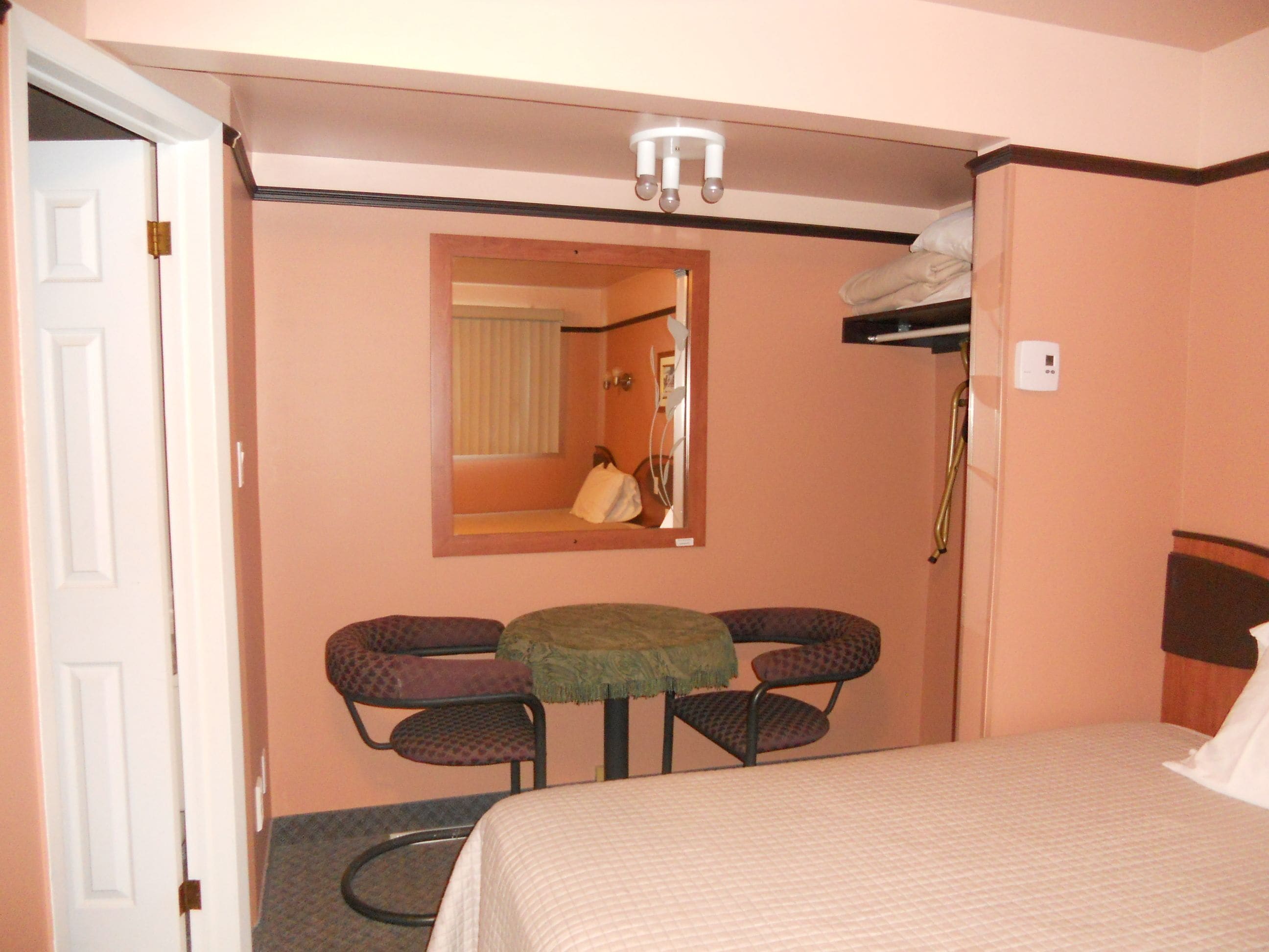 A hotel room with a bed, a small table with two chairs, a mirror, a shower, and a thermostat on the wall.