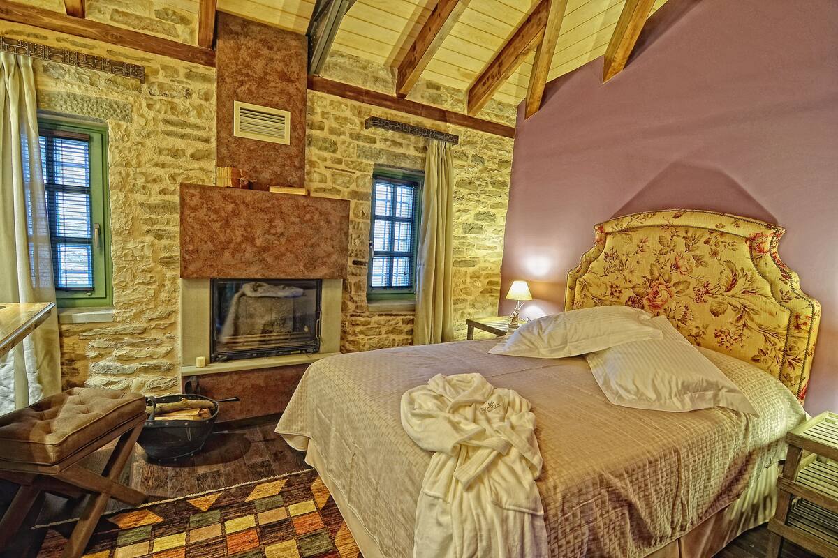 A bedroom with a stone wall, a fireplace, a bed with a floral headboard, and a wooden chair.