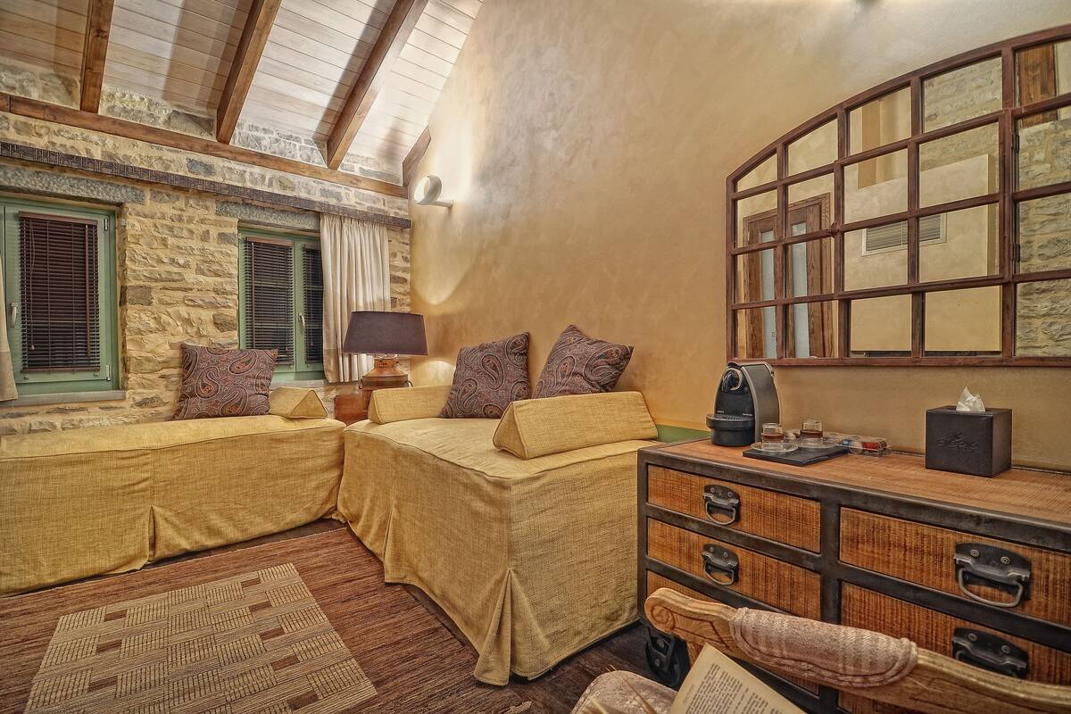 A cozy room with a stone wall, a wooden ceiling, and a large window. There is a bed with a yellow bedspread and two pillows, a wooden dresser, and a lamp on a side table.
