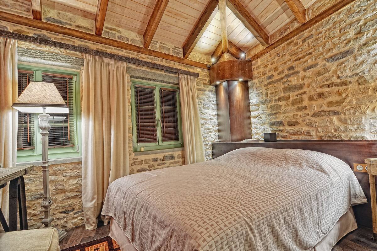 A bedroom with a stone wall, wooden beams, a bed with a patterned bedspread, a nightstand, a lamp, and a window with shutters.