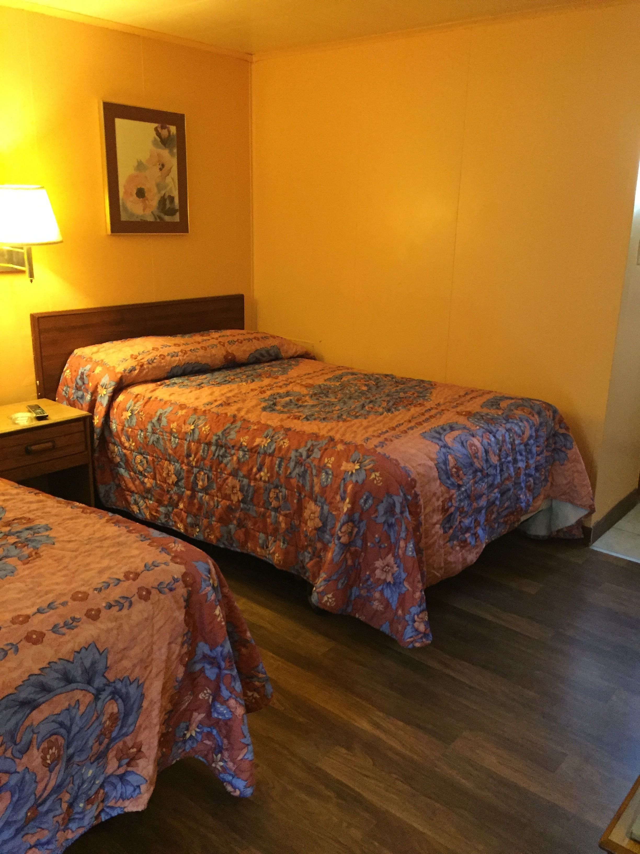 Een hotelkamer met twee bedden, houten vloeren, een nachtkastje met lamp en een ingelijst schilderij aan de muur.