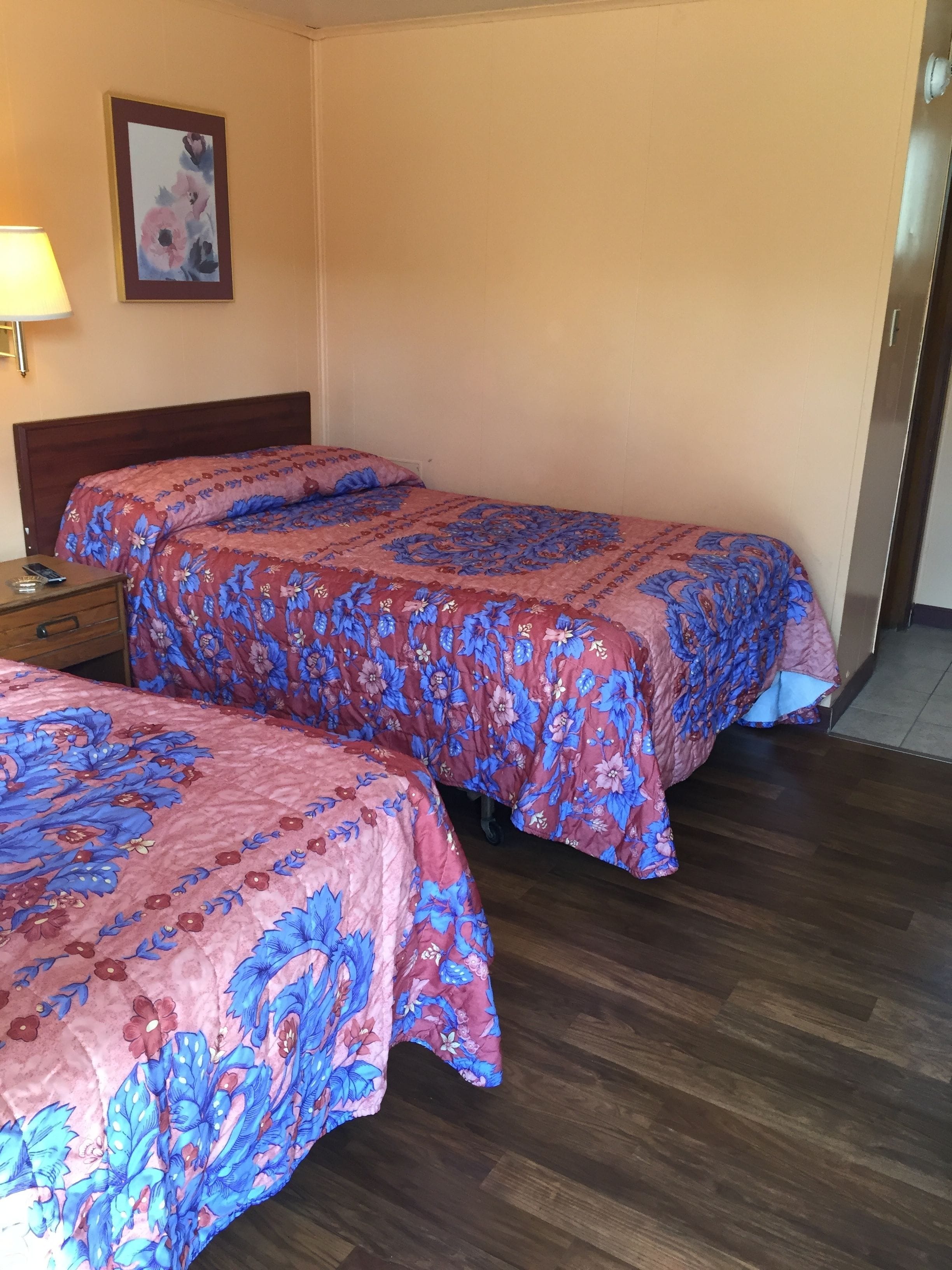 Dos camas individuales con ropa de cama floral, una mesita de noche de madera, una lámpara y un cuadro en la pared.