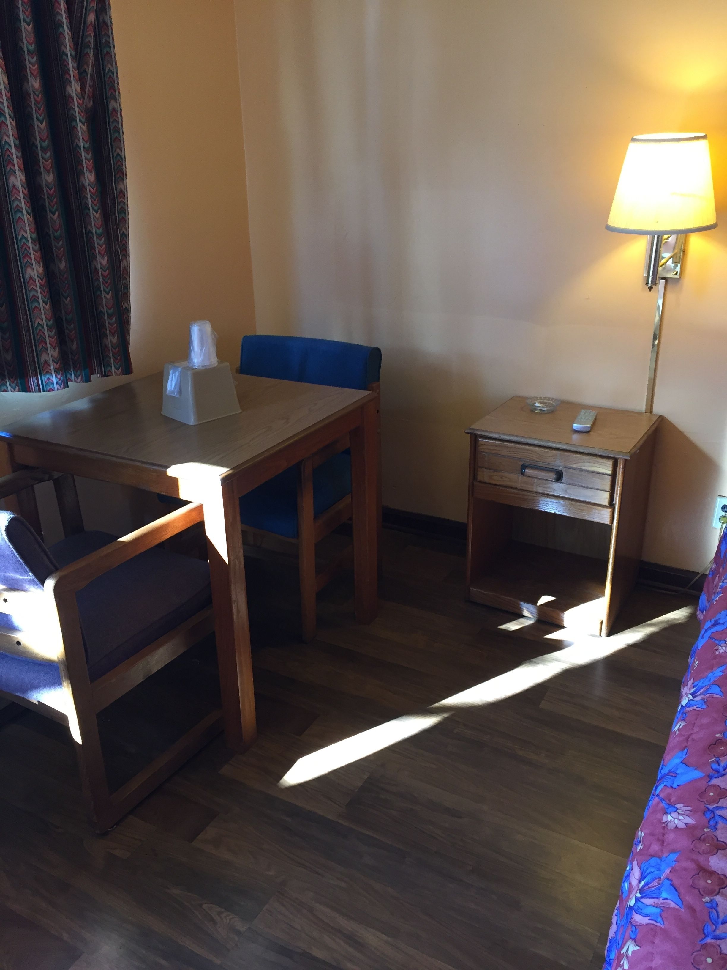 Habitación de hotel con una mesa de madera, una silla azul, mesita de noche con lámpara y una cama con colcha estampada.