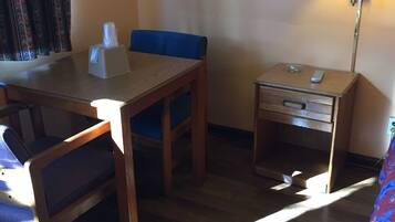 A hotel room with a wooden table, blue chair, bedside table with a lamp, and a bed with a floral bedspread.