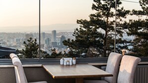Comedor con mesa y asientos acolchados, con vista a un paisaje urbano que incluye edificios y árboles.