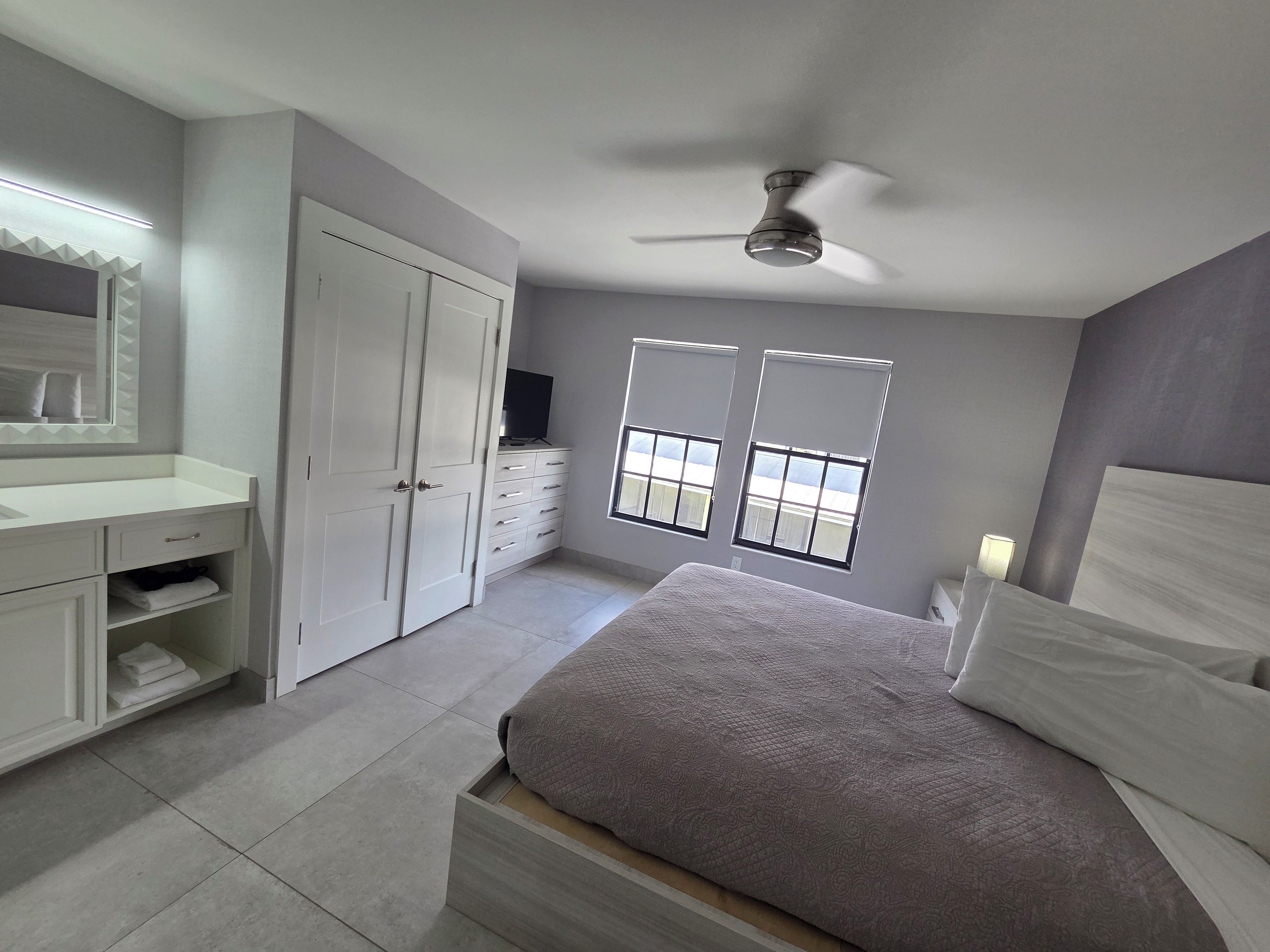 A bedroom with a bed, a wardrobe, a vanity with a mirror, and two windows.