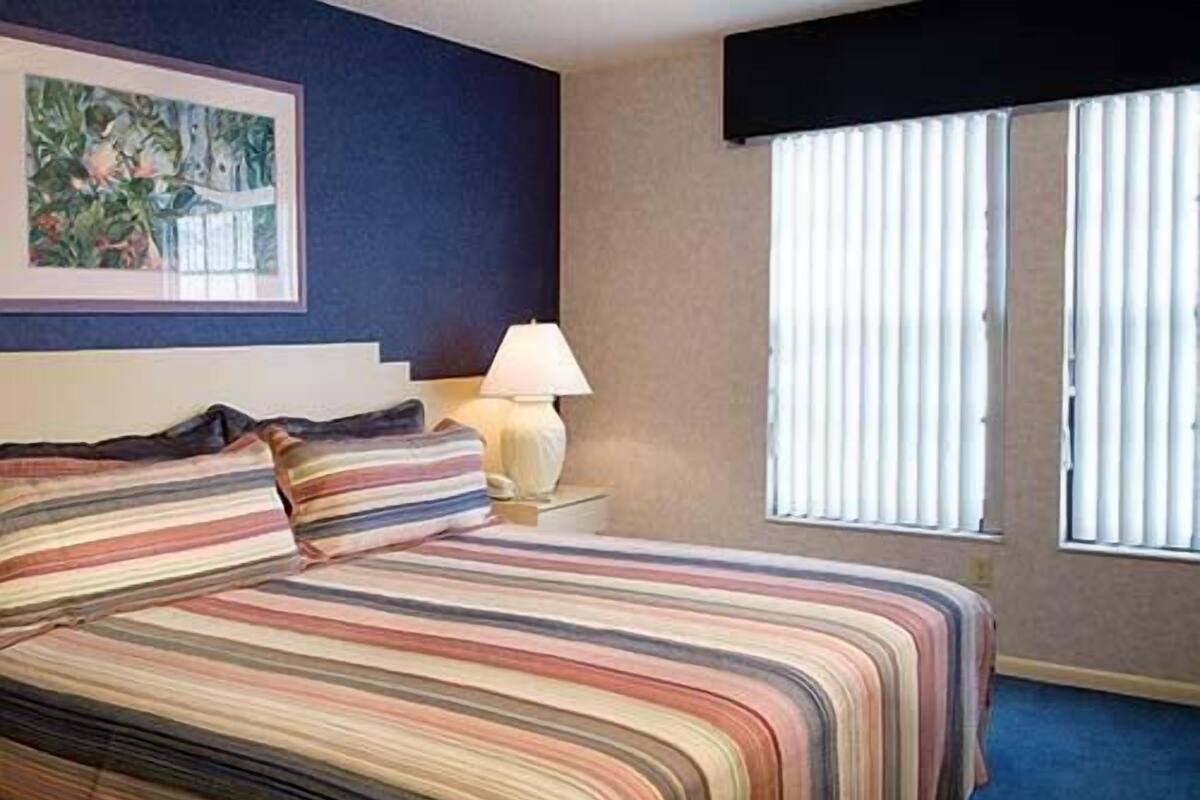A hotel room with a bed, a nightstand, a lamp, a framed picture on the wall, and a window with blinds.