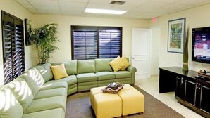 A living room with a green sectional sofa, a yellow ottoman, a flat-screen TV, a plant, and a window with blinds. - High Point World Resort (Kissimmee)