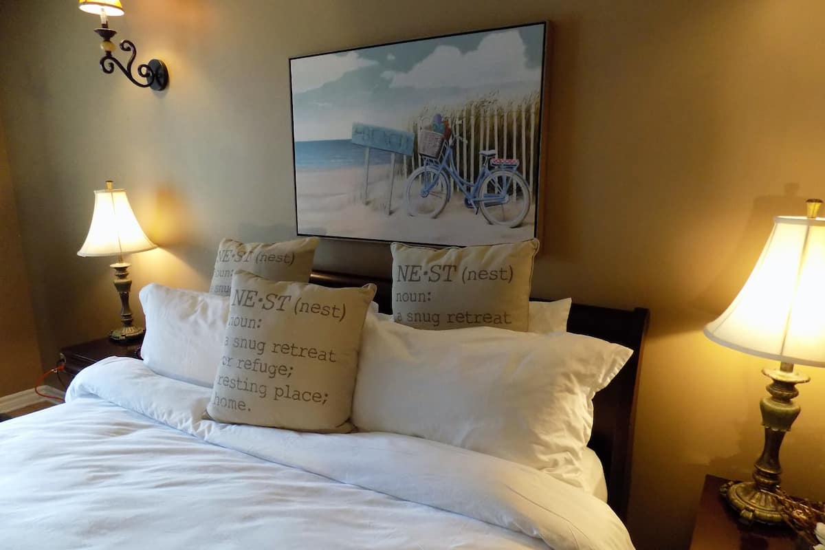 A bed with white bedding, two bedside lamps, and decorative pillows with text.