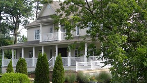 A two-story house with a porch, surrounded by greenery and a stone feature. - L'Oasis de l'Île, Refuge Urbain (Saint-Eustache)