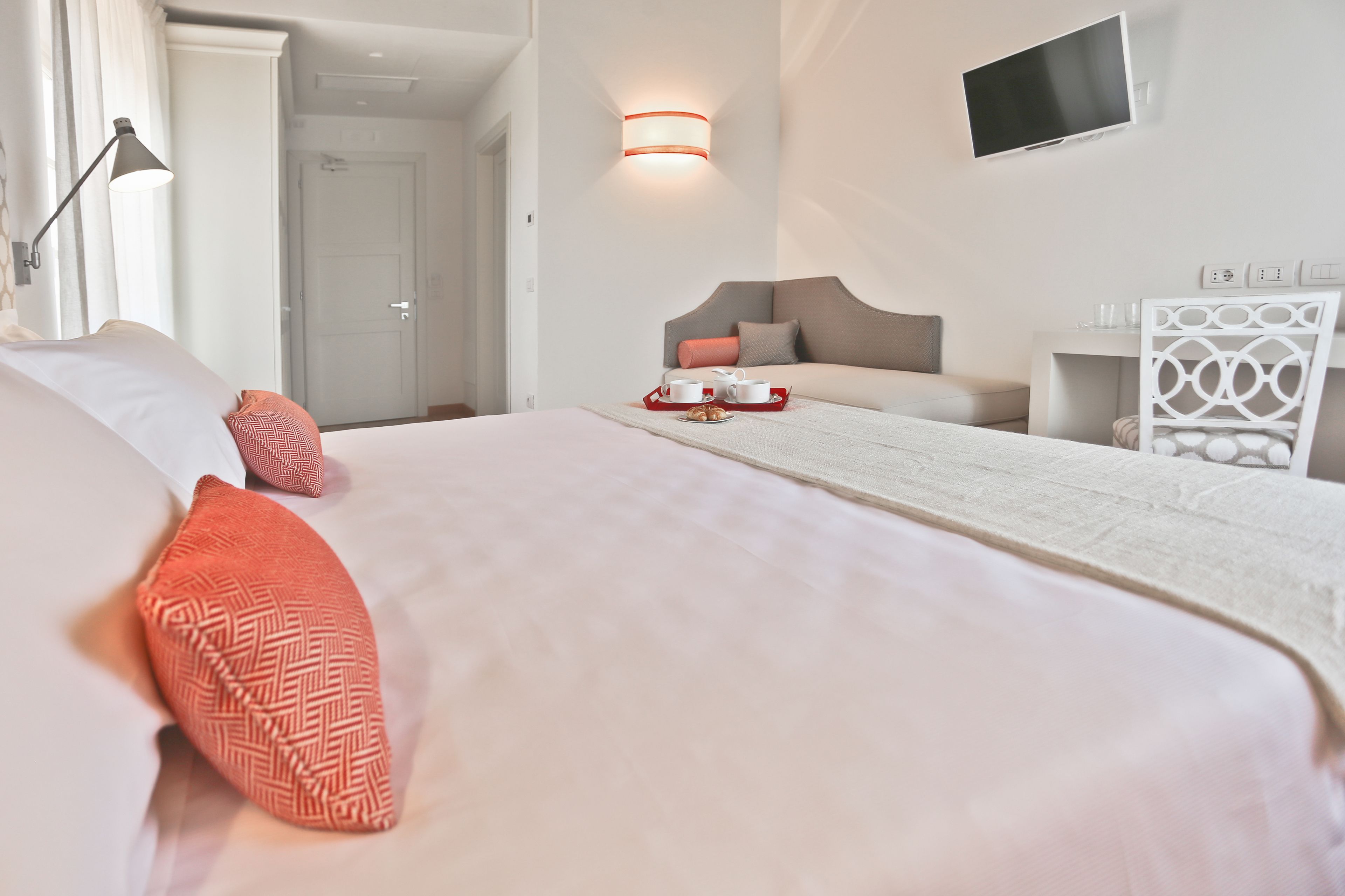 Hotelkamer met een groot bed, een nachtkastje met een dienblad ontbijtartikelen, een aan de muur bevestigde televisie en een witte decoratieve stoel.