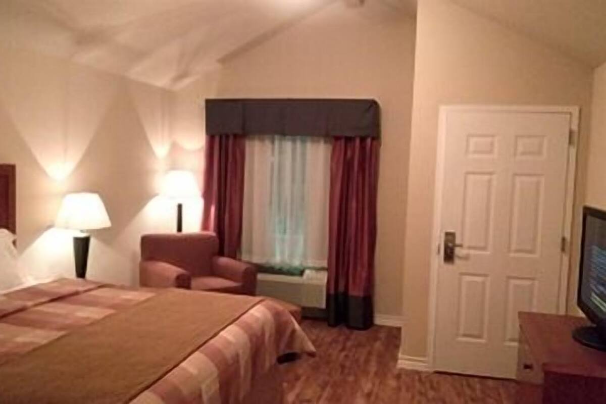 Quarto de hotel com cama, cadeira, escrivaninha com computador, janela com cortinas e porta.