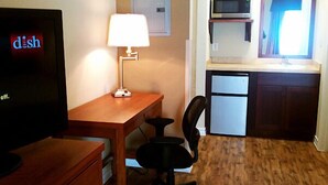 Habitació senzilla amb escriptori de fusta, una cadira, televisió, làmpada, petita cuineta amb microones i nevera, i finestra amb cortines.