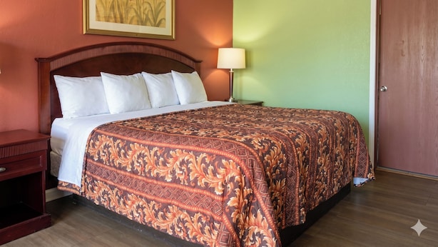 A hotel room with a bed, a nightstand, a lamp, a door, and a painting on the wall. - Budget Inn Jennings LA I-10 (Jennings)