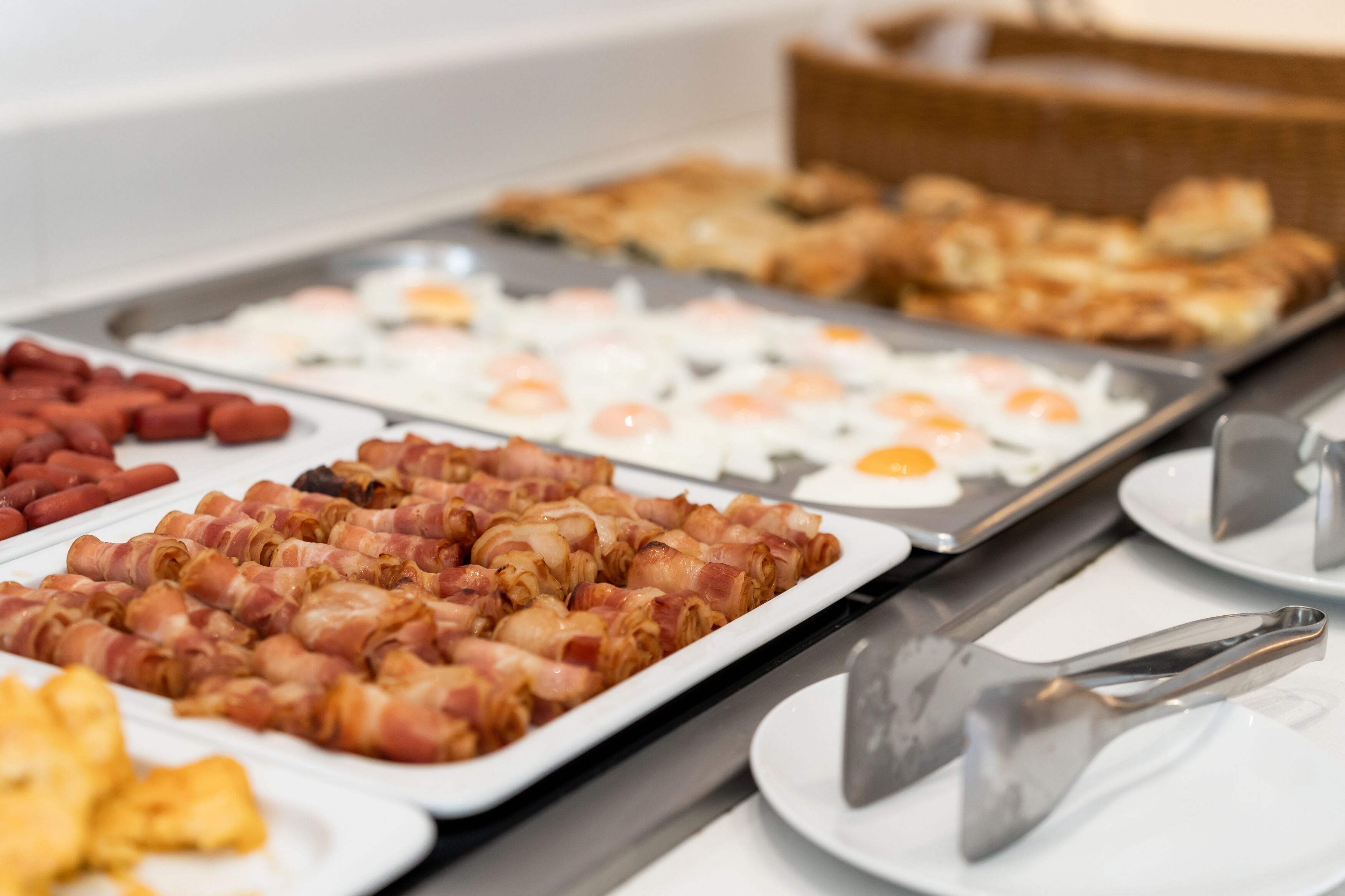 Un bufé con diversas opciones de desayuno, incluyendo salchichas envueltas en tocino, huevos fritos y hot dogs.