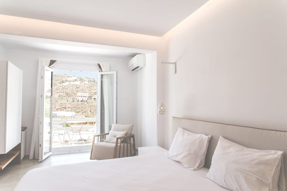 A modern bedroom with a large bed, a chair, and a view of a hillside through an open door.