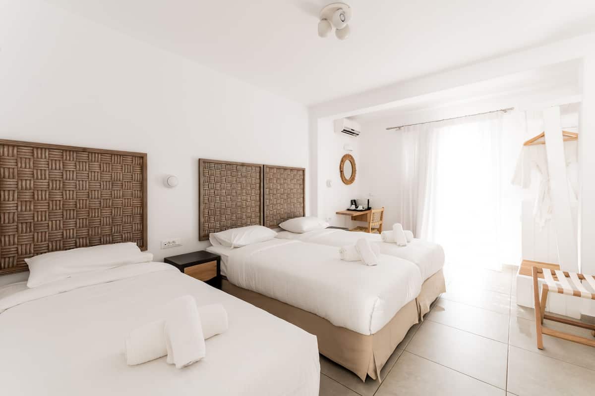 A hotel room with three beds, a wooden headboard, a bedside table with a lamp, and a window with sheer curtains.