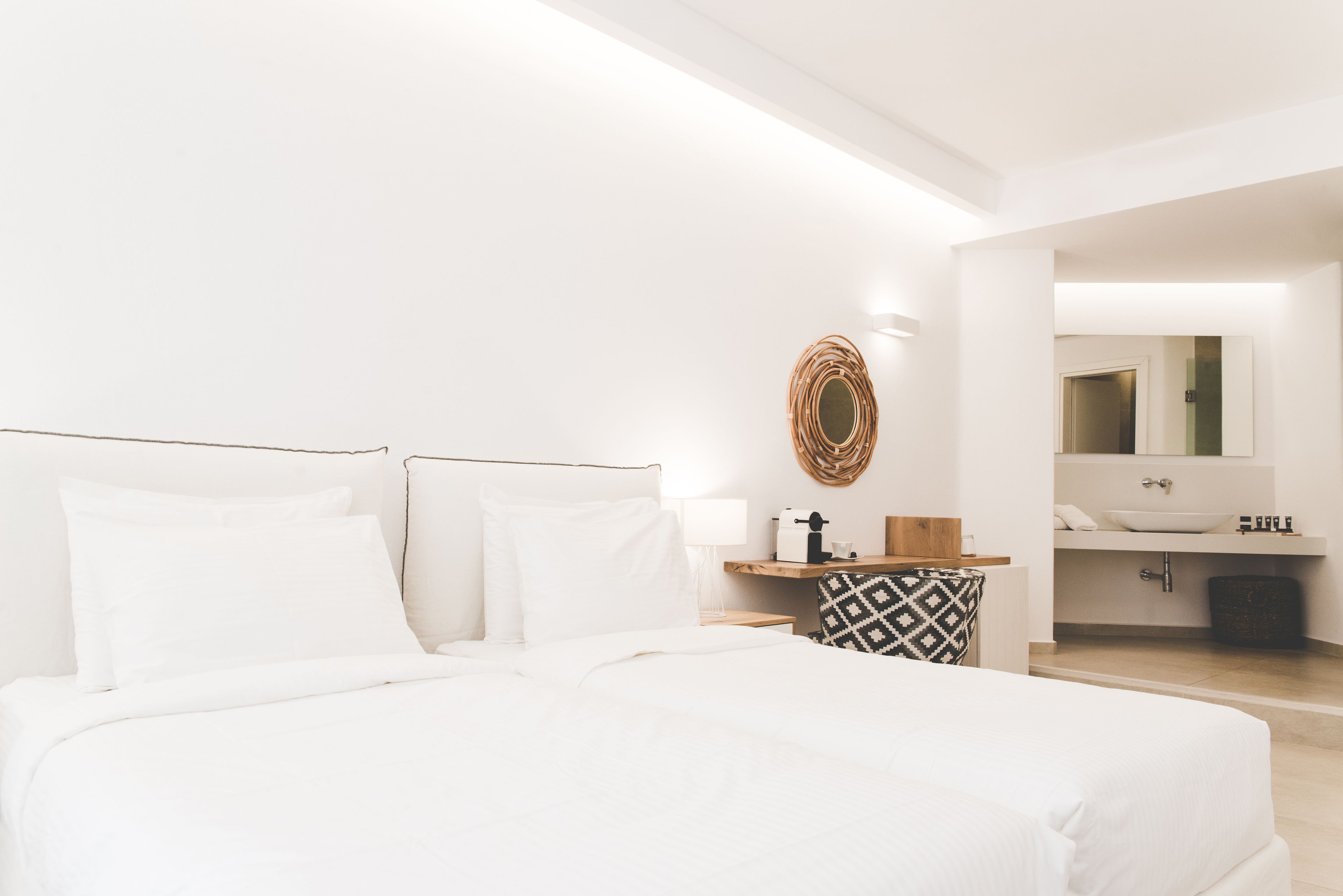 Una habitación de hotel moderna con una cama grande, una mesita de noche con lámpara, una pequeña mesita auxiliar de madera, una silla con estampado y un baño con lavabo y espejo.