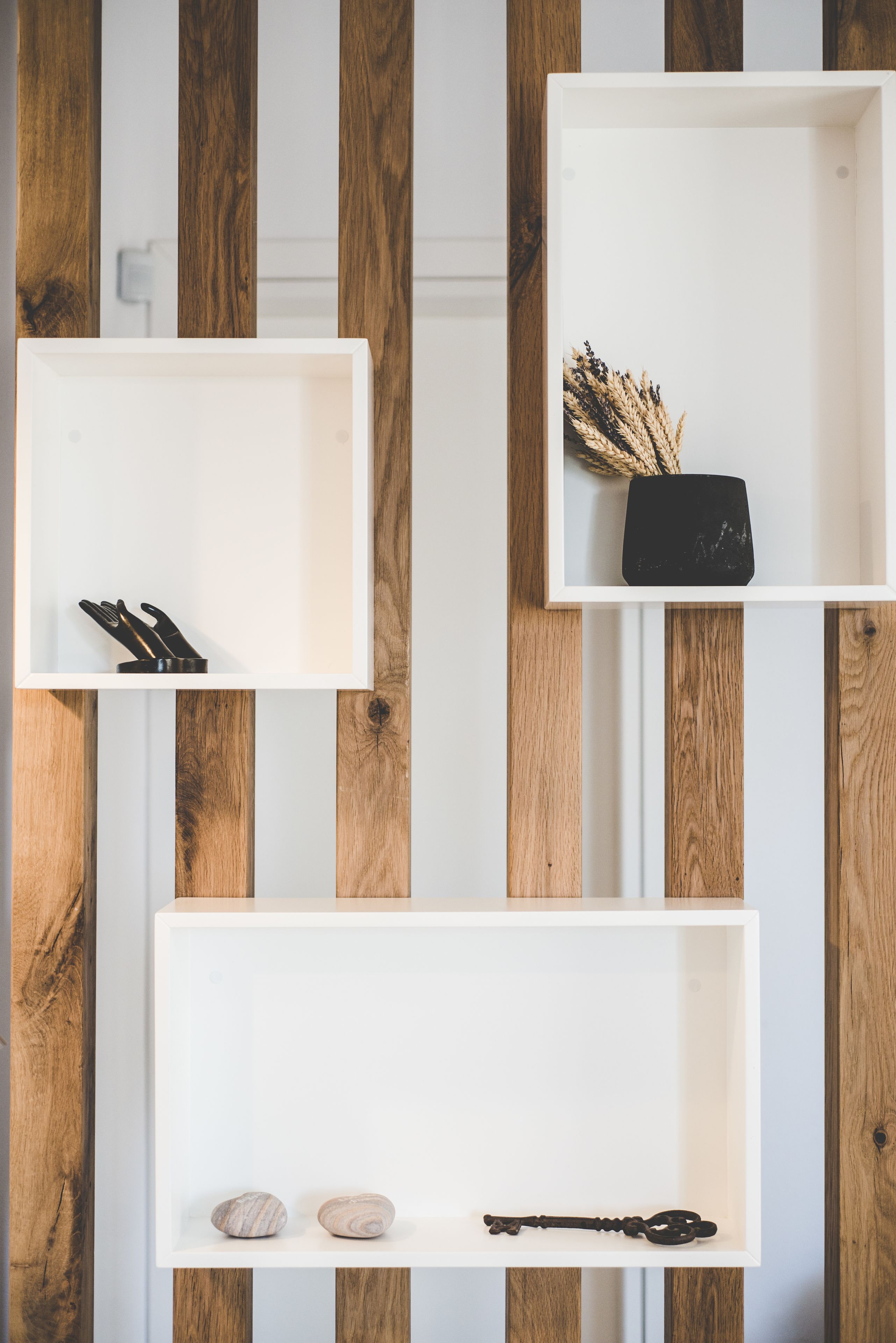 Three white shelves against a wooden wall, each containing different items: the top shelf has a black object and dried plants, the middle shelf has two stones and a key, and the bottom shelf has two stones.