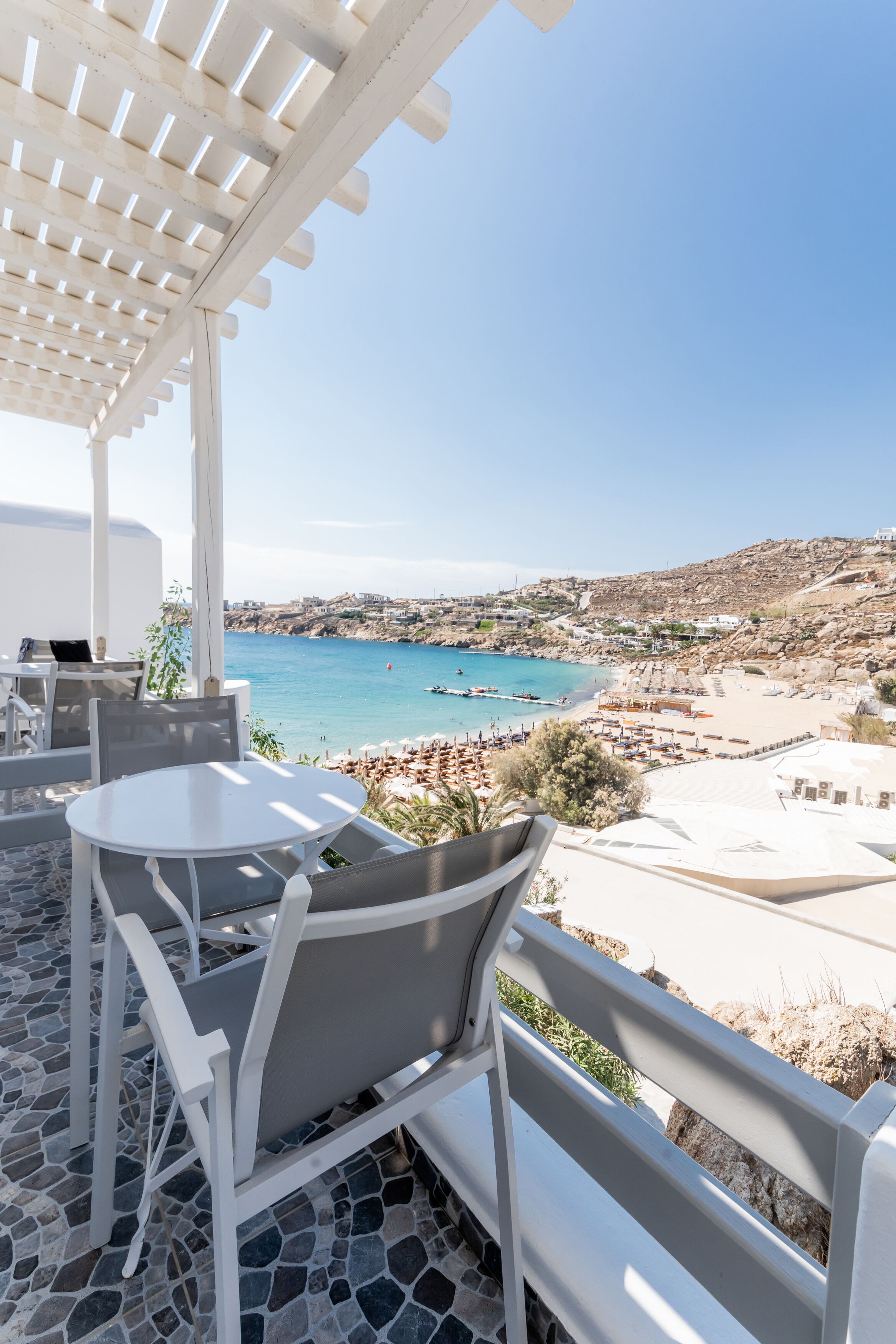 Una terrazza con tavolo e sedie bianche, affacciata su una spiaggia e un paesaggio roccioso.