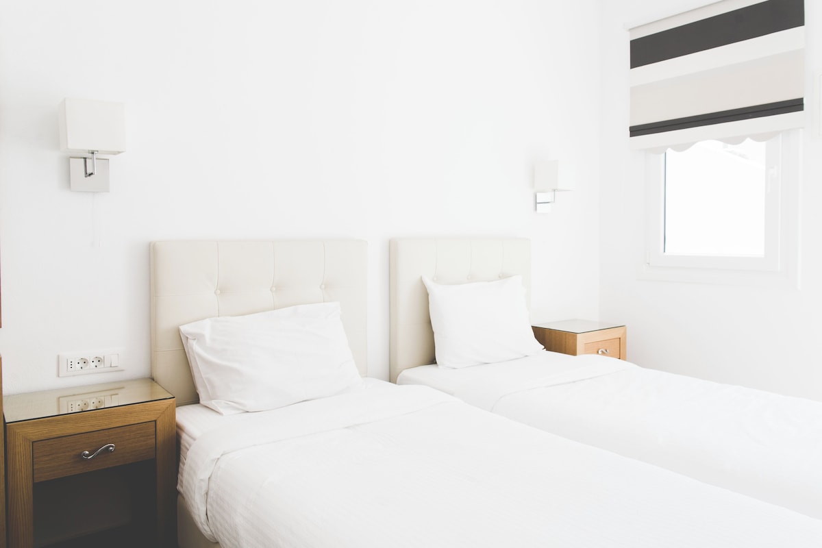 A hotel room with two beds, bedside tables, a wall socket, and a window with blinds.