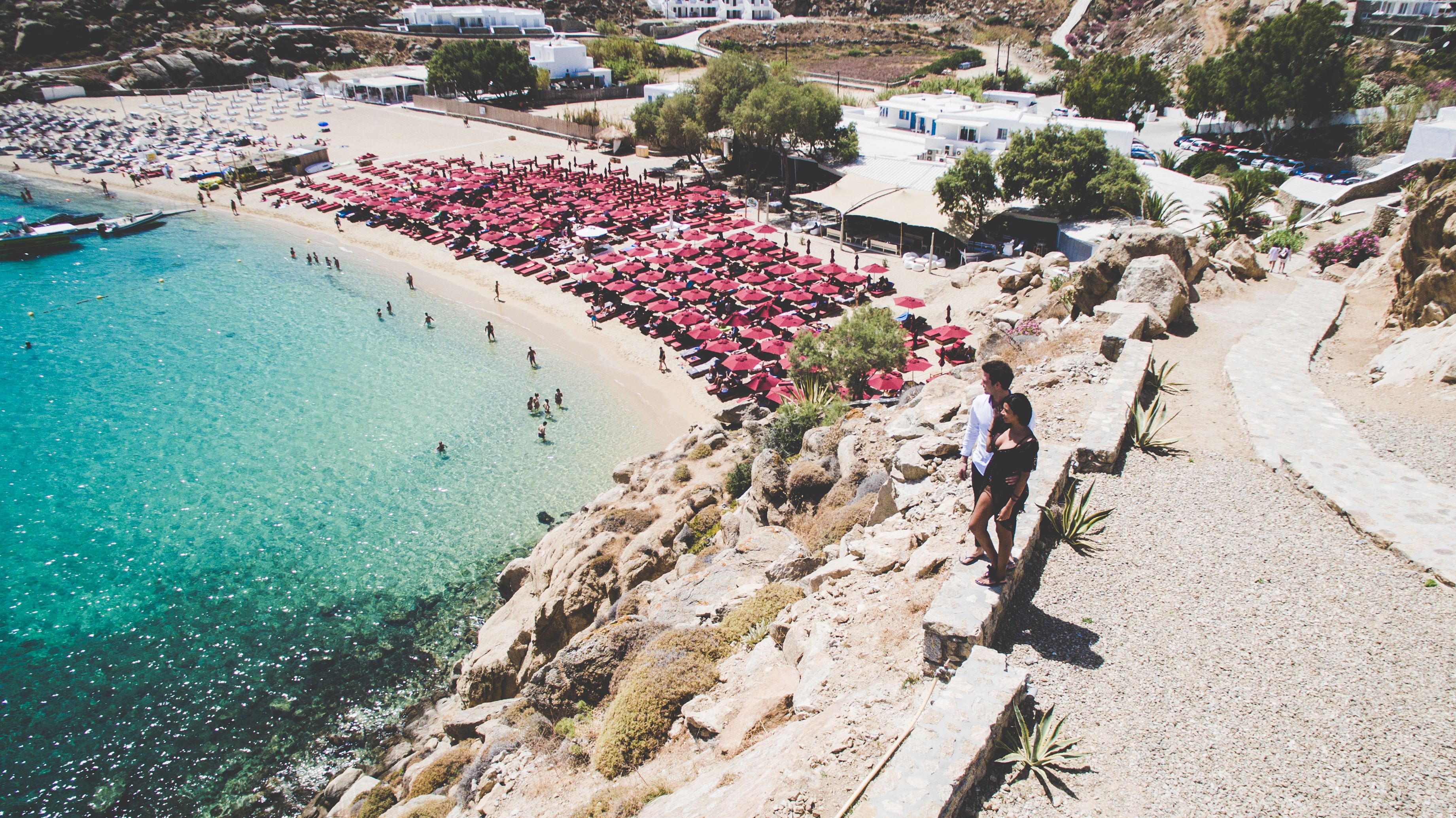 A coastal hotel with a beach view, multiple red umbrellas, and a rocky shoreline.