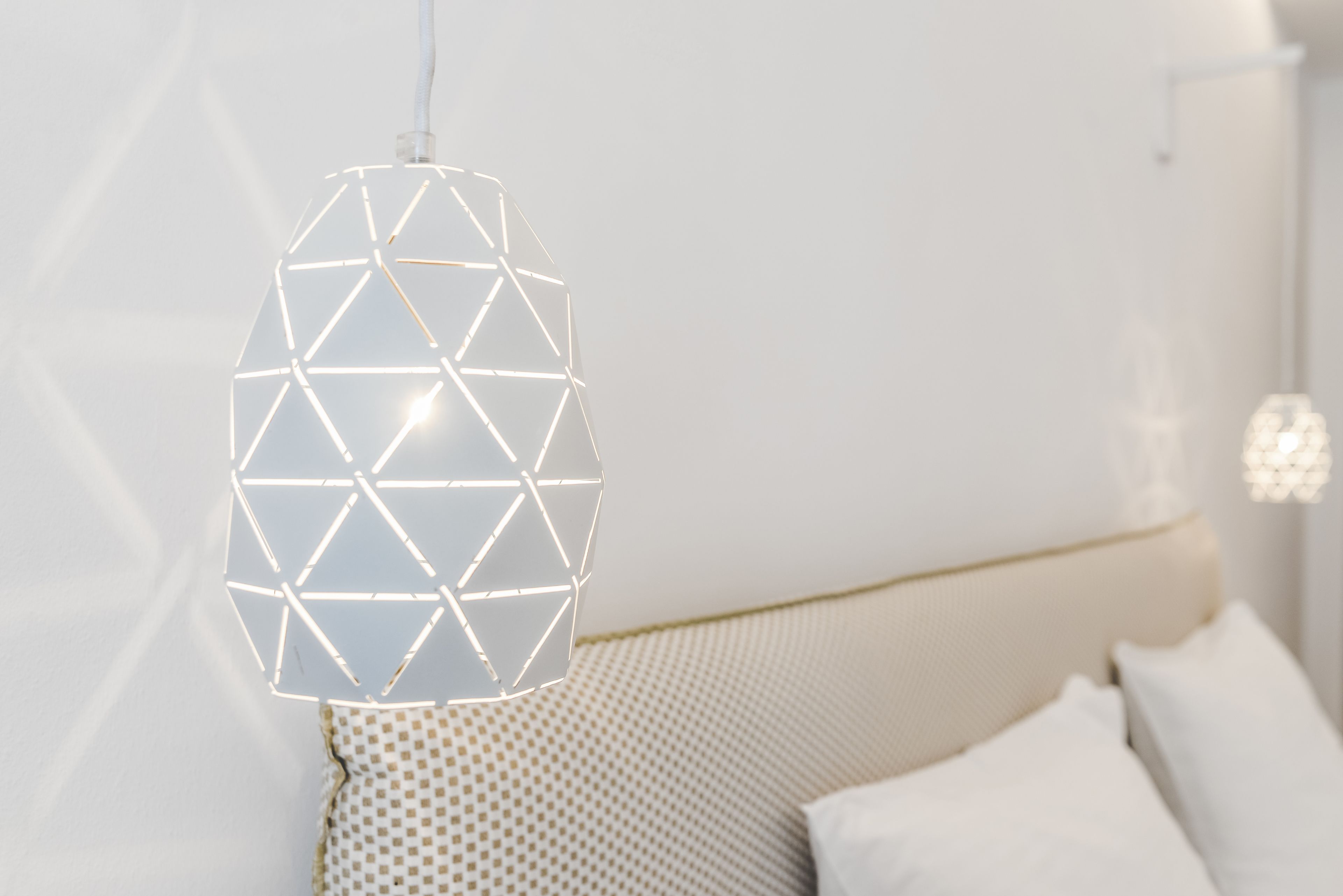 Un luminaire suspendu moderne, de forme géométrique, est installé au-dessus d’un lit bien rangé, doté d’une tête de lit à motifs.