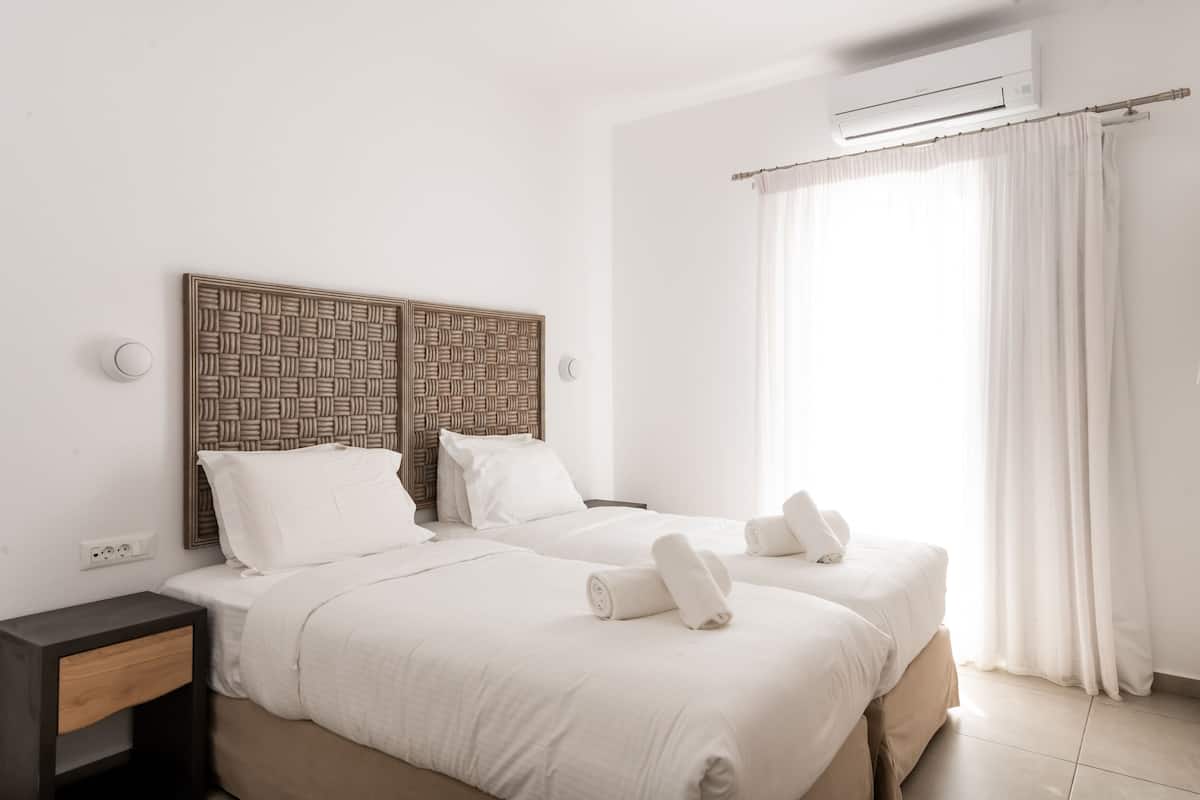 A hotel room with two beds, a woven headboard, white bedding, a nightstand, an air conditioning unit, and a large window with white curtains.