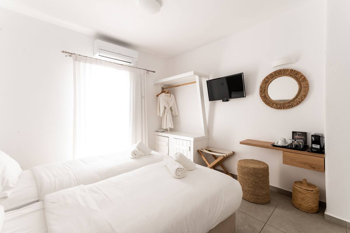 A hotel room with two beds, a TV, a mirror, a shelf with a coffee maker, and a window with curtains.