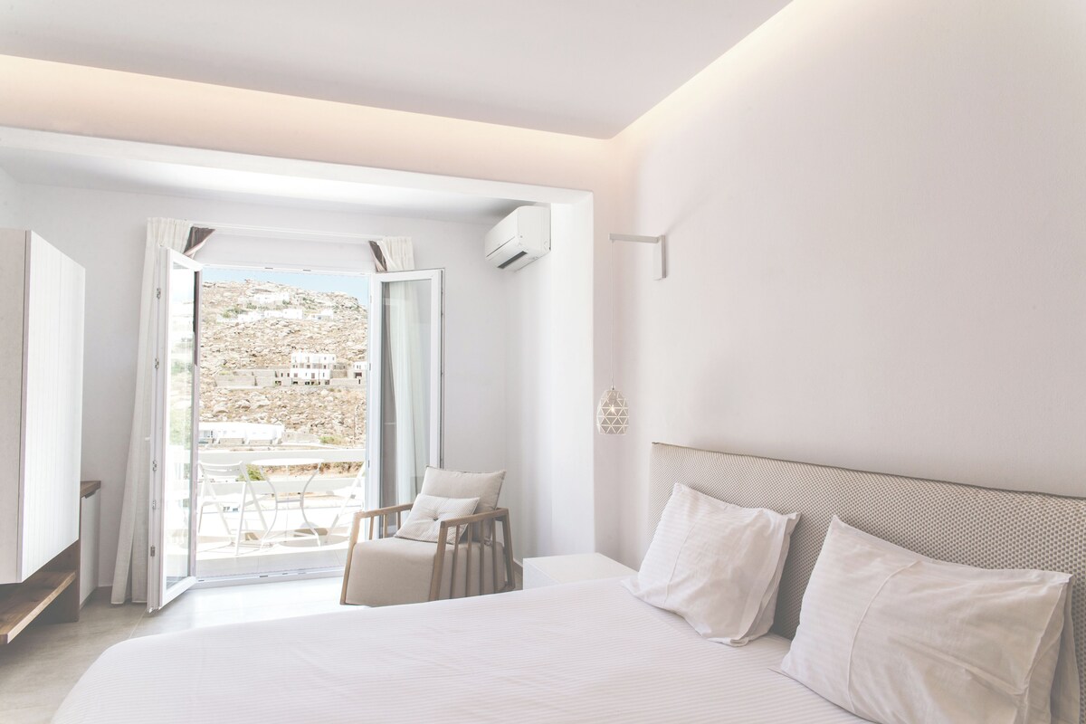 A modern bedroom with a large bed, a chair, and a view of a hillside through an open door.