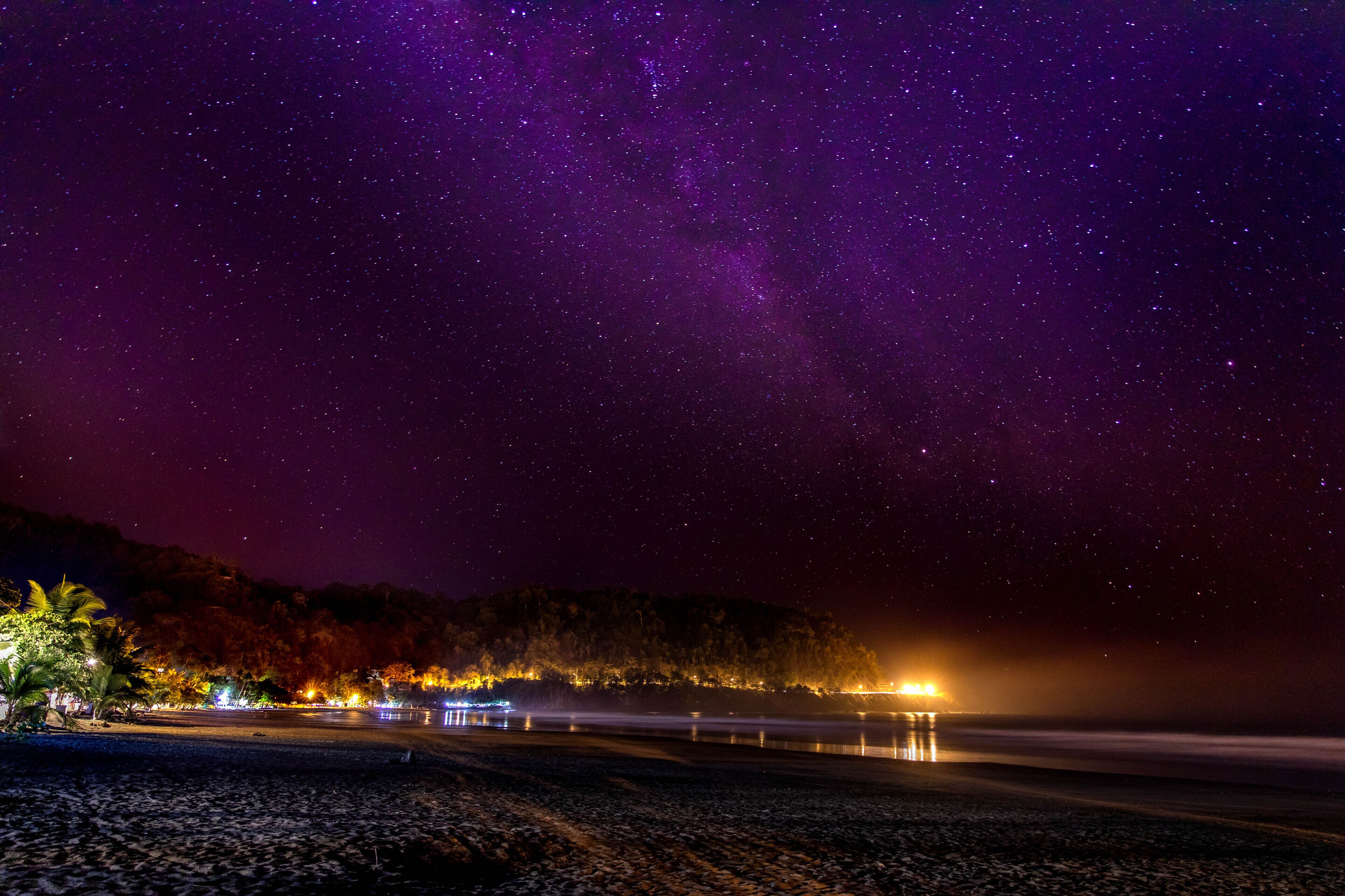 A night scene with a starry sky, a beach, and a forested hill with lights.