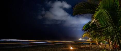 Una playa de noche con una fogata, palmeras y luces de la ciudad a lo lejos.