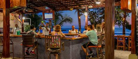 Un bar junto a la playa con estructura de madera, muebles de mimbre y vista al océano.